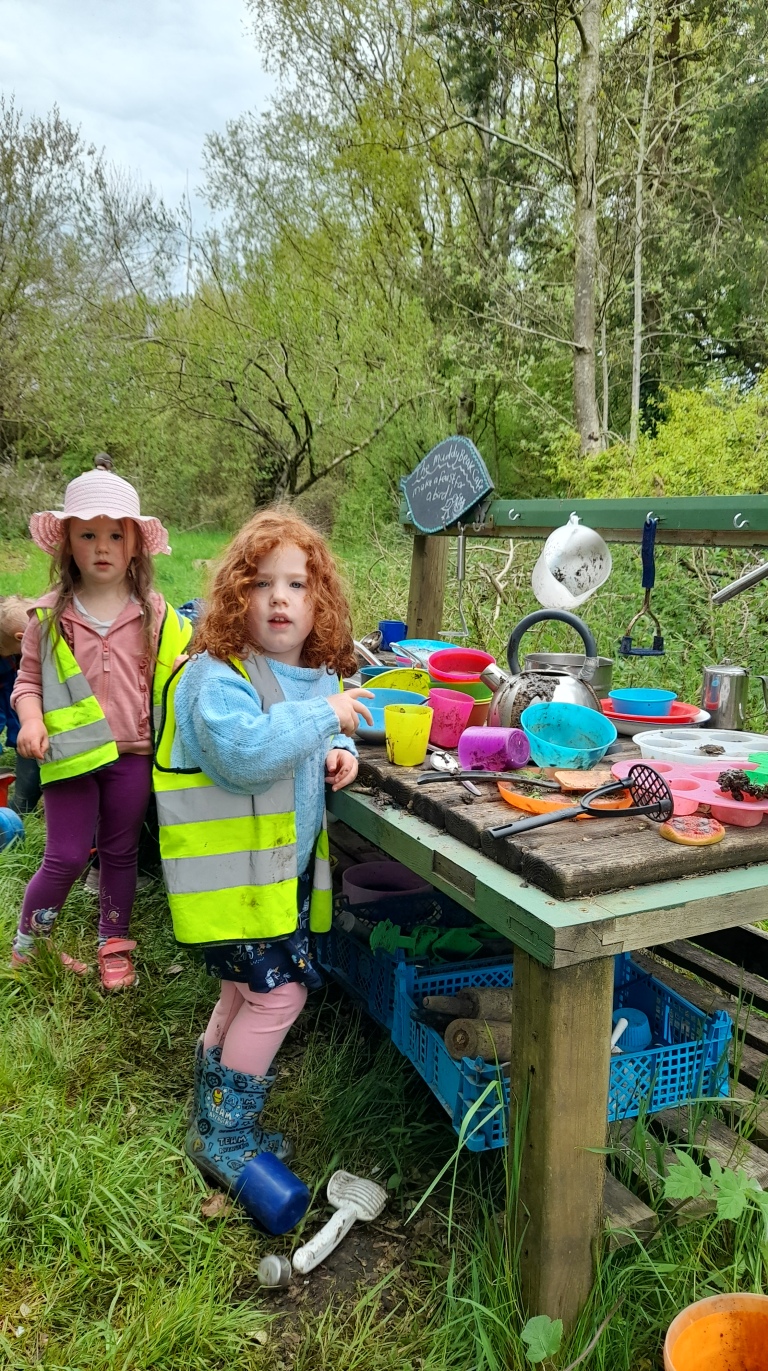Two little girls one with light brown hair and one with ginger curls playing in the mud kitchen